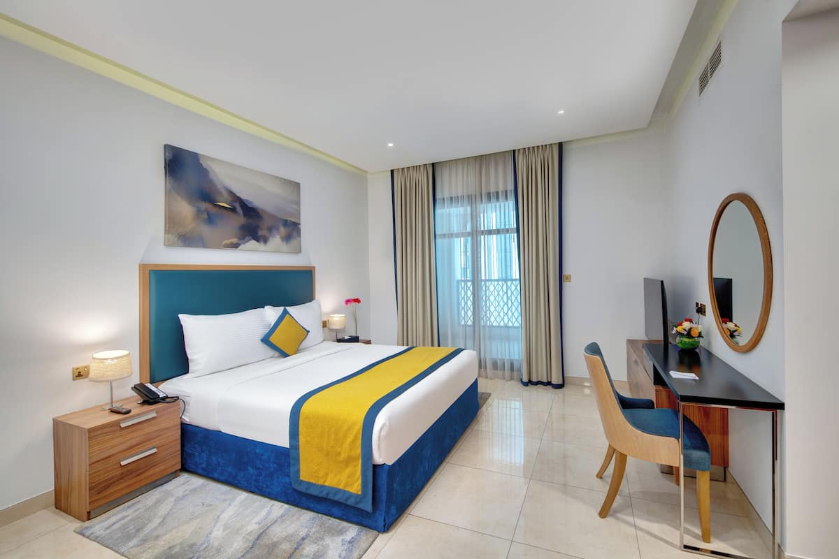 Una habitación de hotel con cama, mesita de noche, escritorio, silla y un amplio ventanal con cortinas.