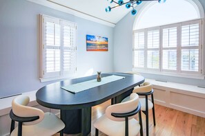 A dining area with a dark wooden table, white chairs, a painting on the wall, and large windows.