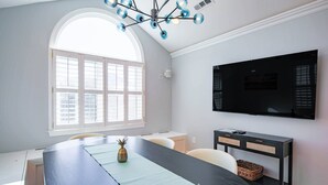 Ein modernes Esszimmer mit einem großen Tisch, einem an der Wand befestigten Fernseher und einem Fenster mit Jalousien.