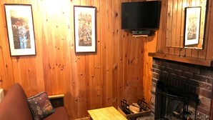 Een gezellige kamer met een houten wand, een flatscreen televisie boven een open haard, een houten tafel en een bruine bank met een kussen met dessin.