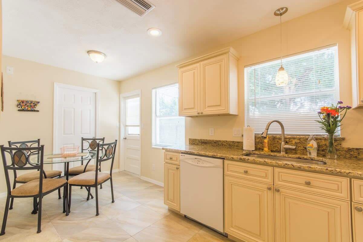 A kitchen with a glass-top dining table, chairs, a dishwasher, cabinets, and a window with blinds.