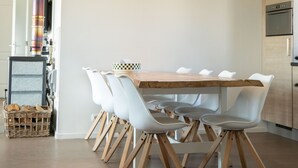 Un coin repas avec une table en bois et des chaises blanches, une branche décorative ornée de cœurs suspendus, et une cuisine moderne en arrière-plan.