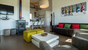 Un salon moderne avec un canapé, un pouf et une table basse, un téléviseur à écran plat, une cheminée et une cuisine en arrière-plan.