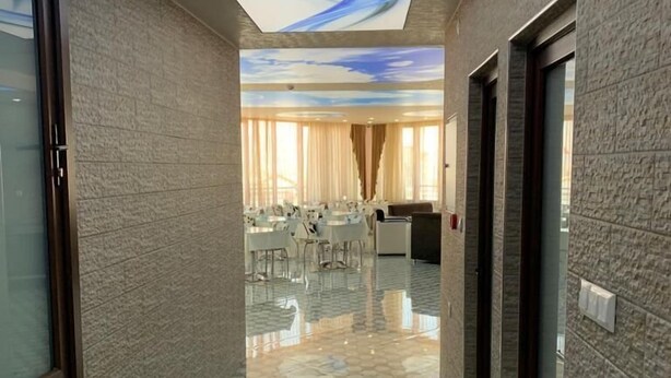 A hallway with a patterned floor, a ceiling with a blue and white design, and a room with tables and chairs in the background.