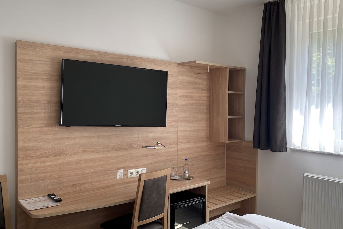 A hotel room with a flat-screen TV mounted on a wooden wall, a desk with a chair, a small refrigerator, and a window with sheer curtains.