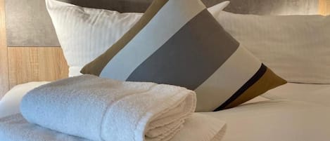 Una cama bien hecha con sábanas blancas y una toalla doblada, junto con un cojín de estampado geométrico.