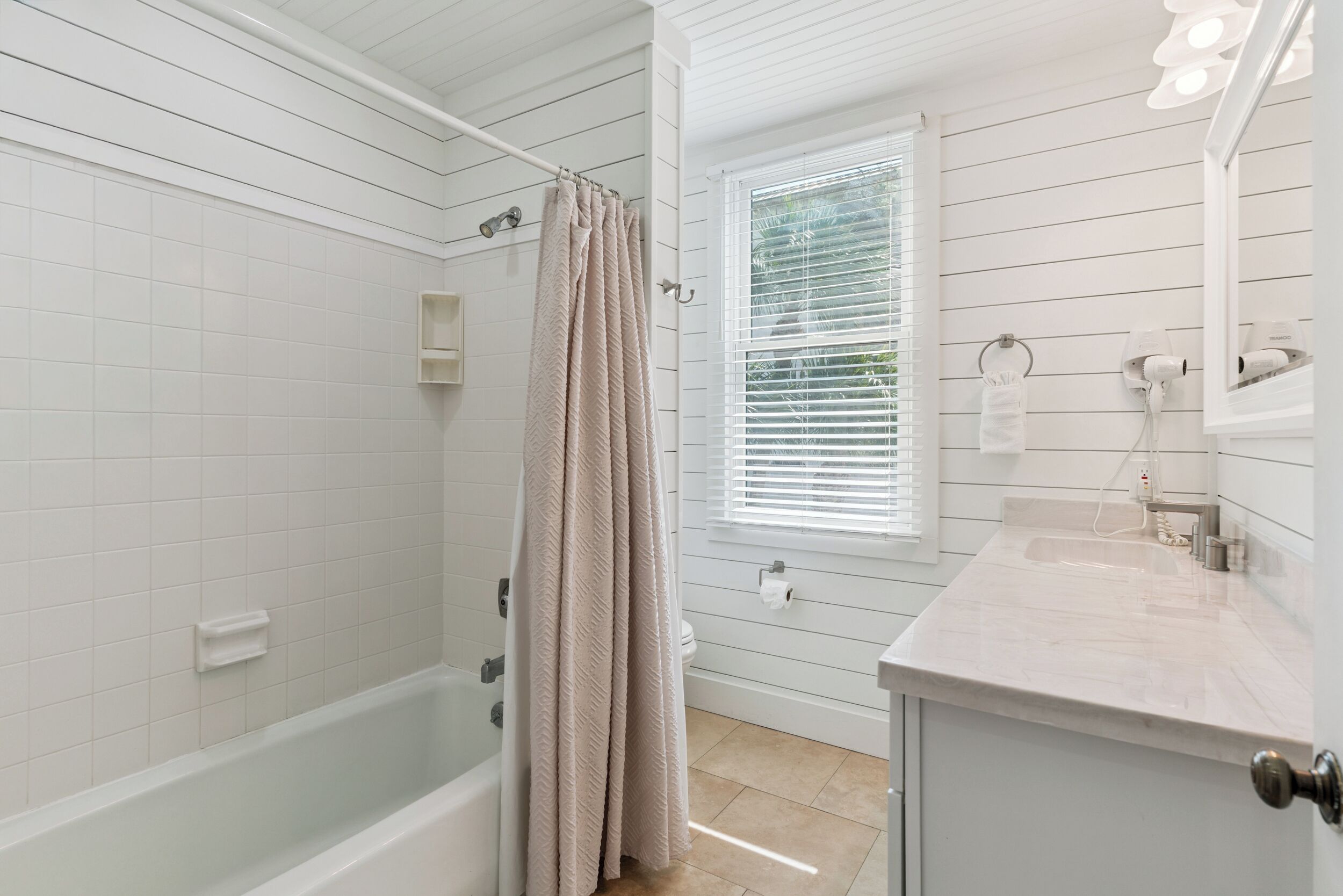 Ein Badezimmer mit Badewanne, Duschvorhang, Fenster mit Jalousien, Waschbecken und Spiegel.