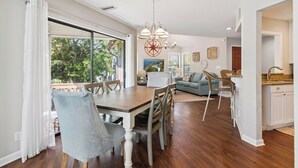 A dining area with a wooden table and chairs, a kitchen with white cabinets, and a living room with a sofa and chairs.
