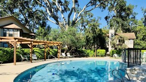 A swimming pool with a wooden pergola and lounge chairs, surrounded by trees and a residential building.
