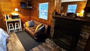 Interior acolhedor de uma cabana com lareira, um sofá com almofadas, uma mesinha de centro com abajur e uma escada.