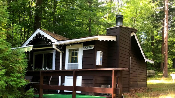 Uma cabana pequena com um deck verde, uma janela e uma chaminé.