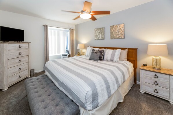 A hotel room with a bed, a TV, a desk, a chair, a ceiling fan, and artwork on the wall.
