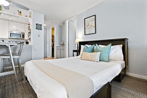 Et hotelværelse med seng, sengebord, lampe og et tekøkken med mikroovn og lille køleskab.