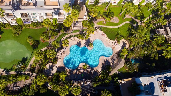 Relax by the wavy pool surrounded by lush greenery and palm trees, with a tennis court and lawn area nearby.