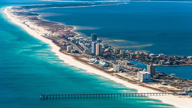 Vista aérea de uma cidade costeira com uma extensa praia arenosa, um píer que se estende sobre a água, e vários edifícios altos ao longo da costa.