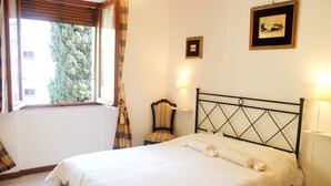 Un llit ben arropat amb capçalera, una cadira, una taula petita i una finestra amb cortines.