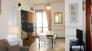 Un saló amb un sofà, una taula de dinar amb cadires, una televisió sobre un moble, i una zona de cuina amb armaris i un frigorífic.