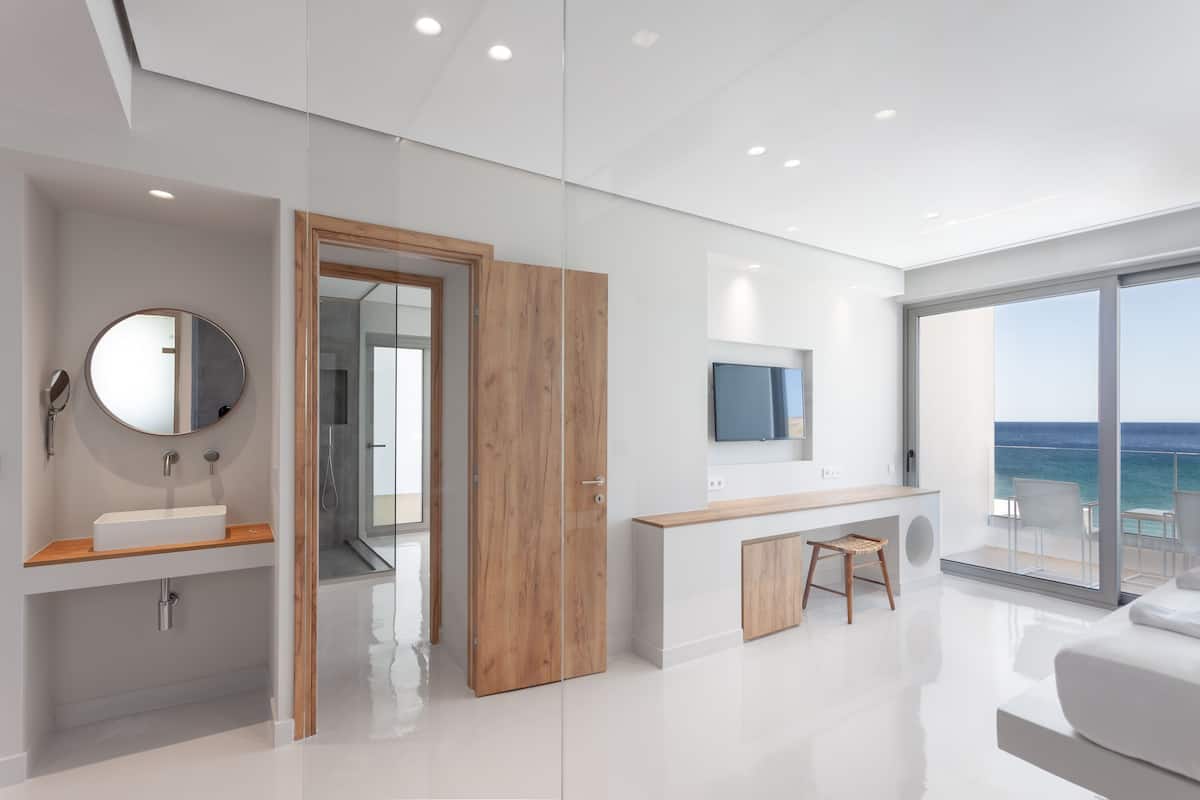 Un dormitorio moderno con un ventanal grande, una puerta de madera, un espejo redondo, un lavabo blanco y un televisor de pantalla plana.