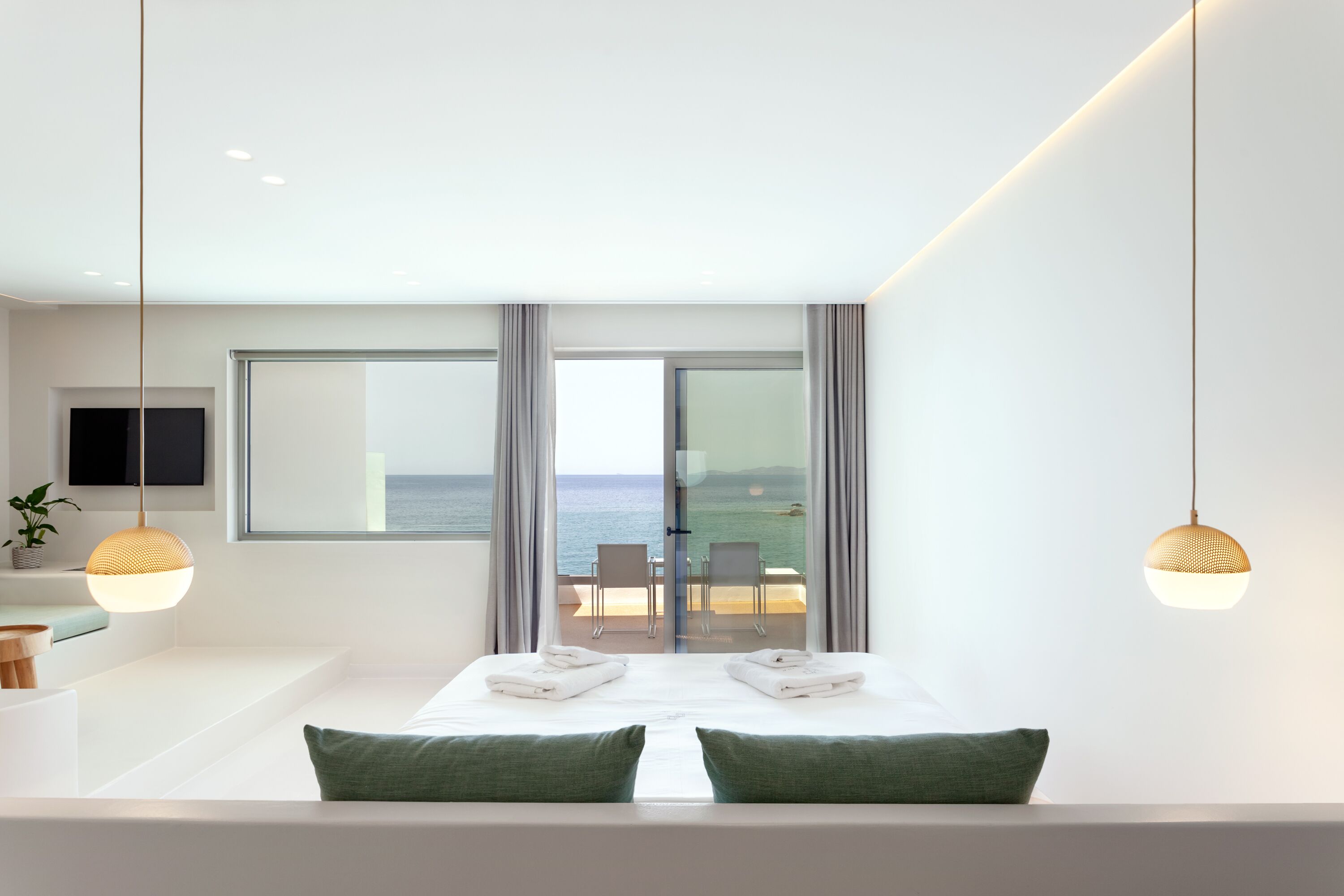 Une chambre d’hôtel moderne avec un grand lit, deux oreillers verts, une télévision et un balcon offrant une vue sur la mer.