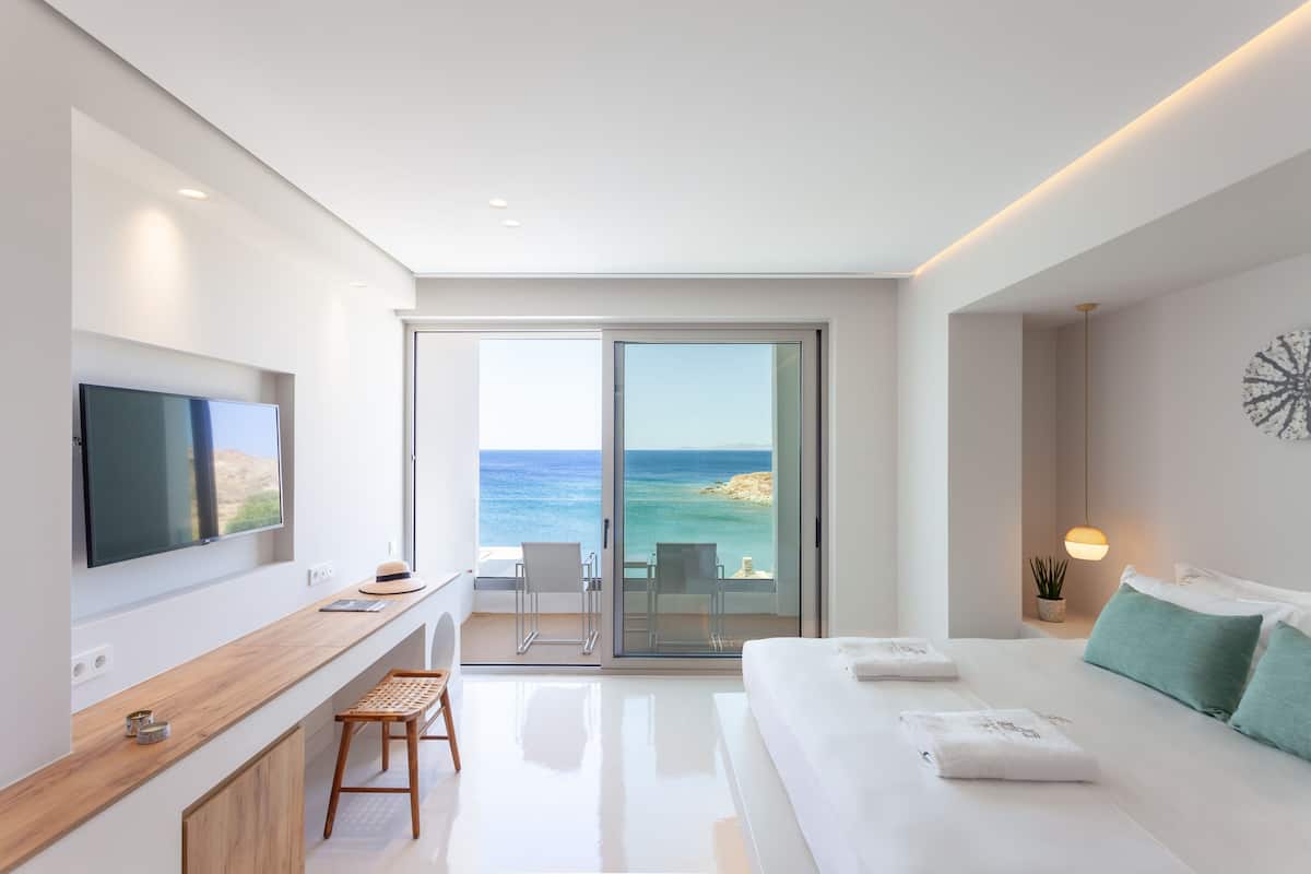 Habitación de hotel moderna con un balcón amplio, televisión de pantalla plana, escritorio de madera y cama con ropa blanca.