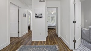 A hallway with wooden flooring, white walls, and a patterned rug. There are doors leading to other rooms, and a ceiling light fixture is visible.