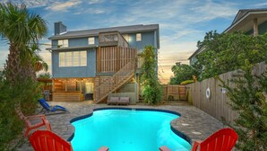 A backyard with a swimming pool, red Adirondack chairs, a wooden deck, and a two-story house with a balcony. - Sand Castle ~ 3-Story Beach Retreat with Heated Pool, Tiki Bar & Dual Game Rooms (Panama City Beach)