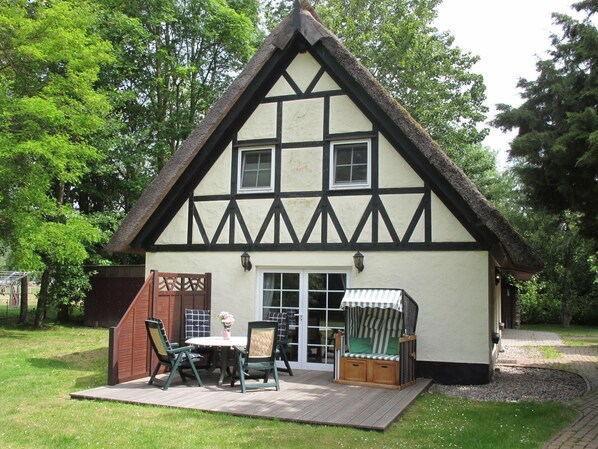 A traditional half-timbered house with a thatched roof, a wooden deck, and a small outdoor seating area with chairs and a table.