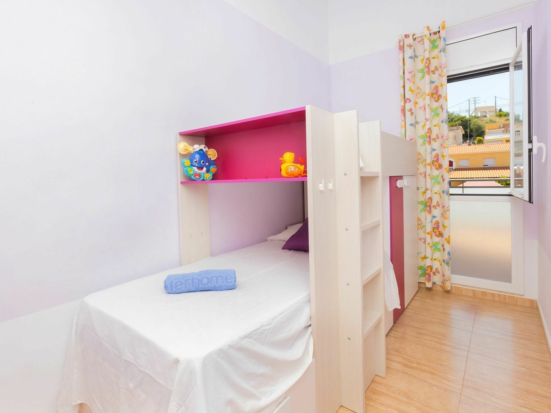 A small, single-bed room with a bunk bed, a white bedspread, a pink shelf with toys, and a window with a floral curtain.