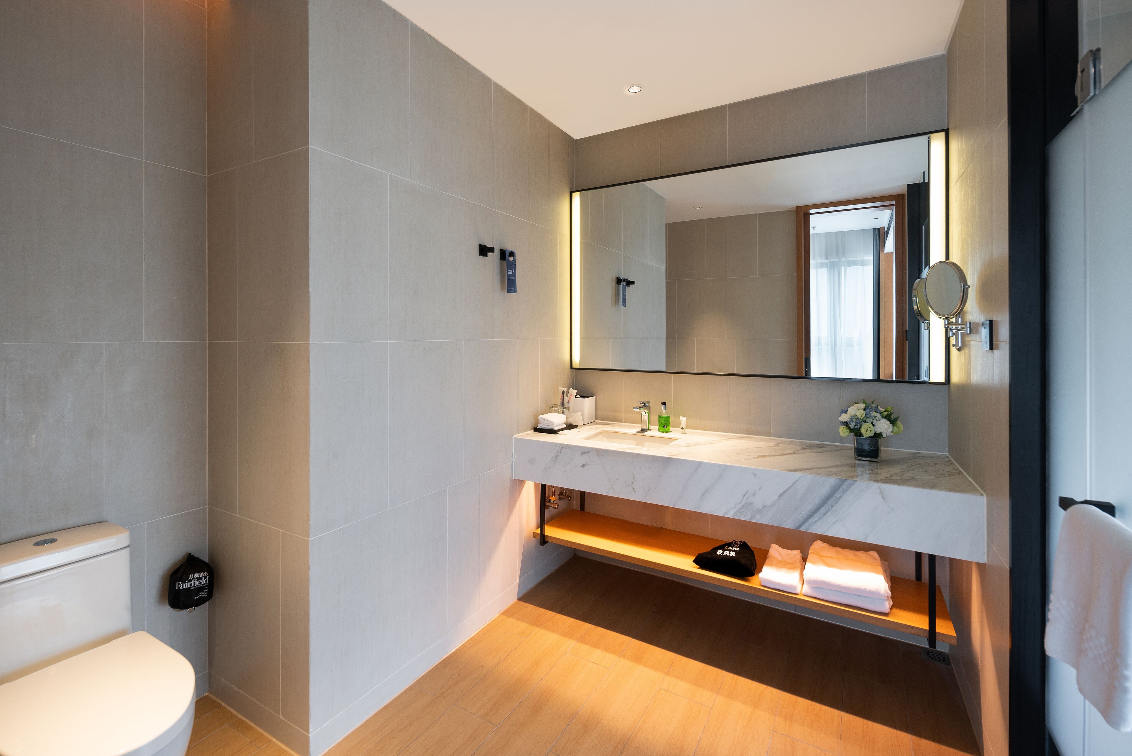 Een moderne badkamer met toilet, wastafel met een marmeren blad, spiegel en een plank met handdoeken.
