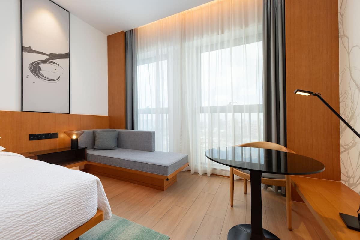 A modern hotel room with a bed, a sofa, a small table, and a large window with curtains.