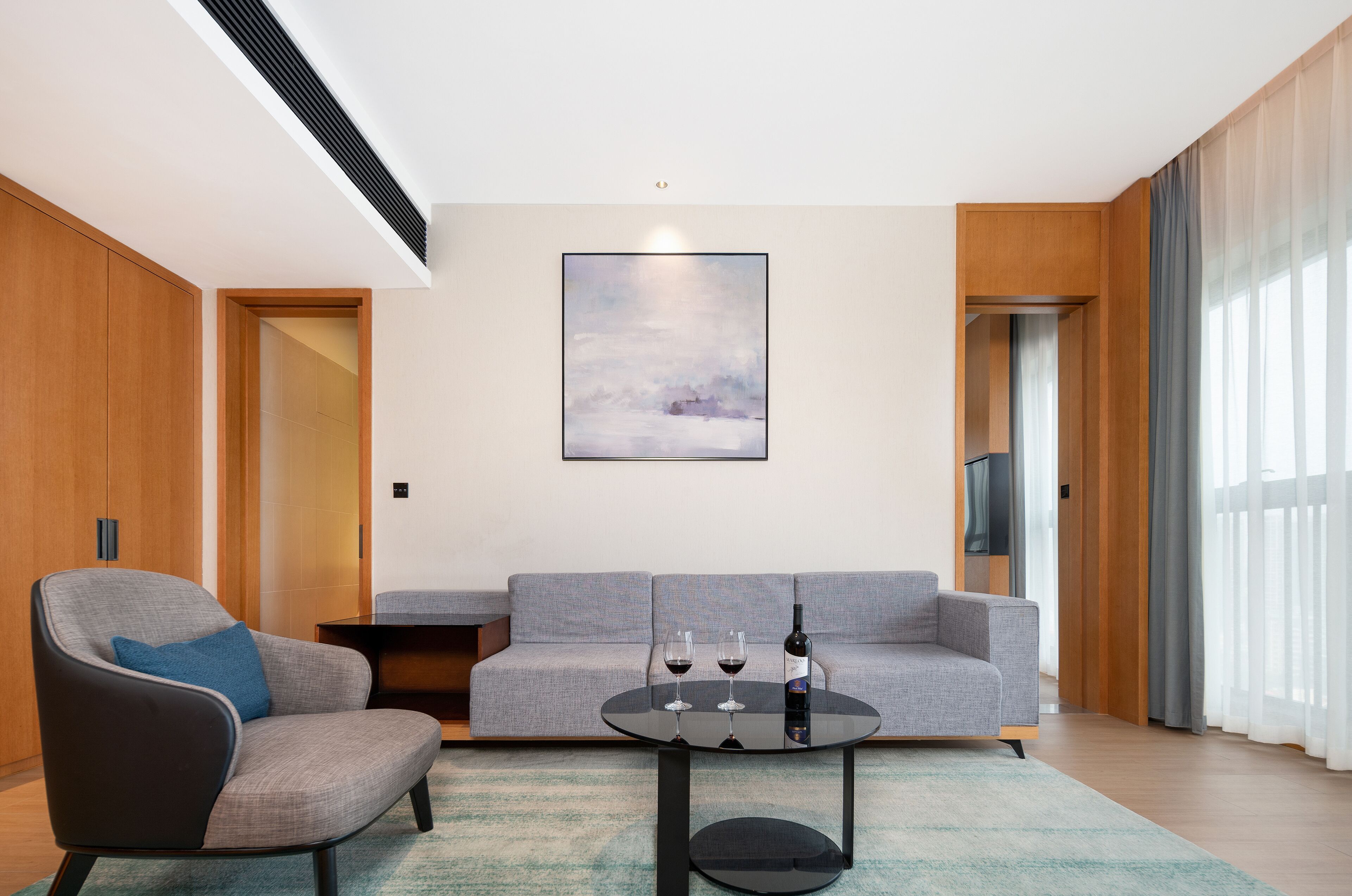 A modern living room with a grey sofa, a black armchair, and a round coffee table. There is a painting on the wall and a television on a wooden stand.