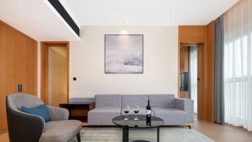 A modern living room with a grey sofa, a black armchair, and a round coffee table. There is a painting on the wall and a television on a wooden stand.
