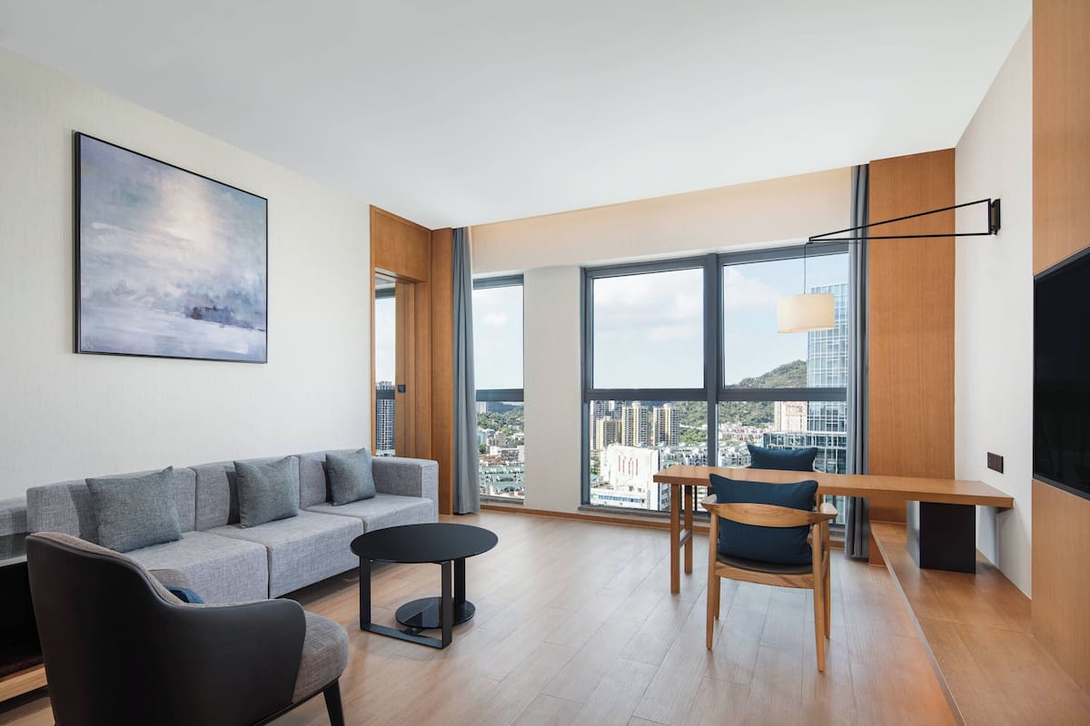 A modern living room with a grey sofa, a black armchair, a small round table, and a wooden bench. Large windows offer a view of the cityscape.