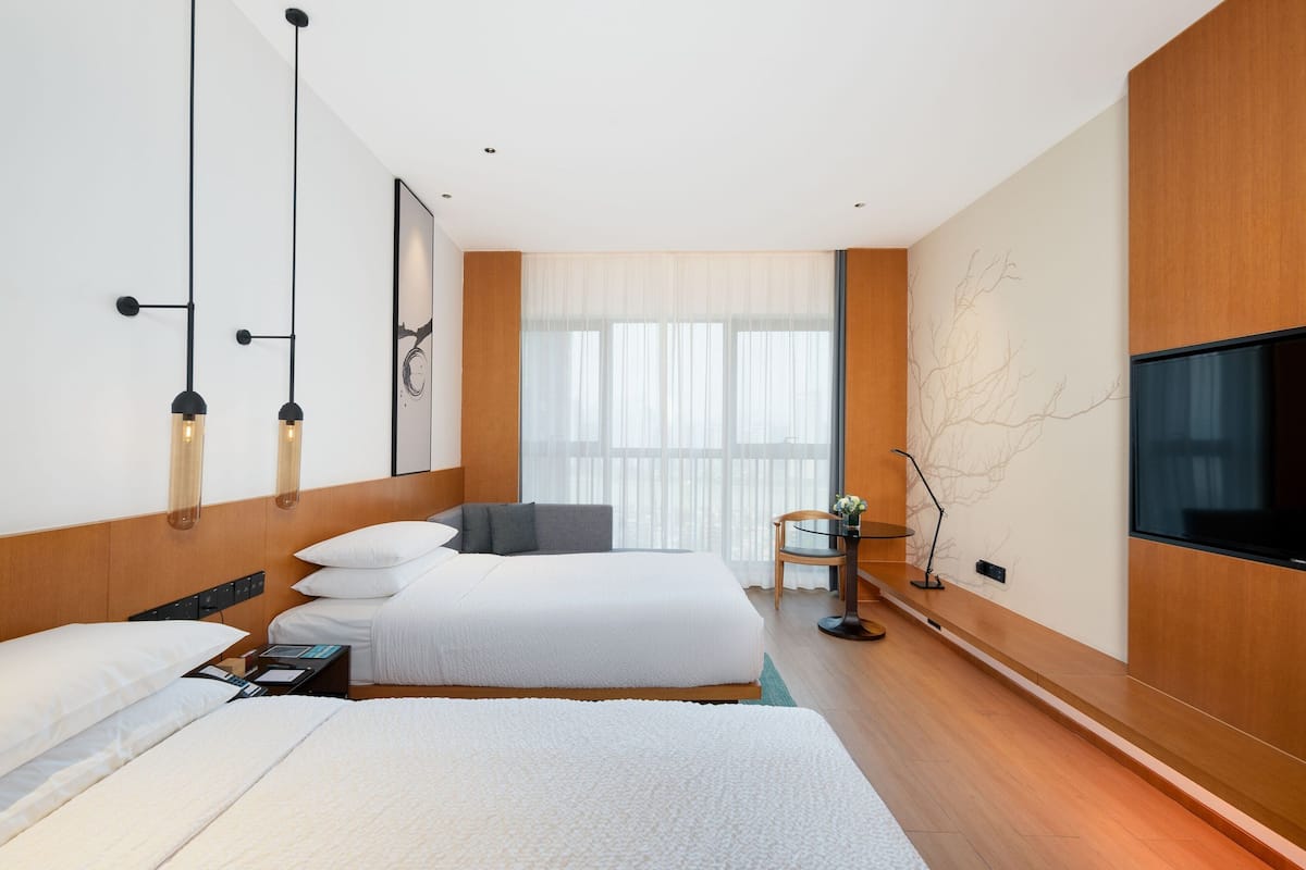 A modern hotel room with two beds, a flat-screen TV, a small table, and a large window with sheer curtains.