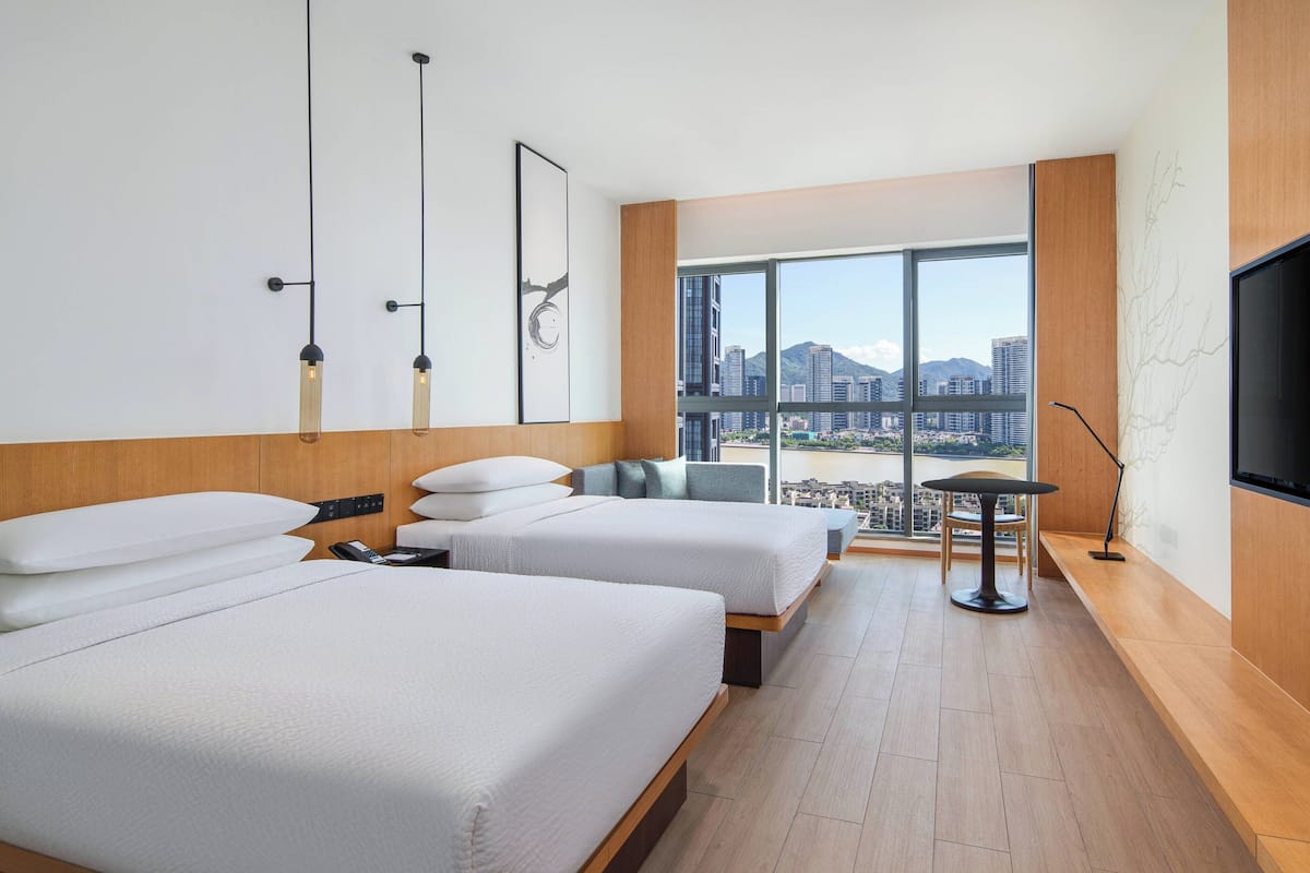 A modern hotel room with two beds, a desk, a chair, and a large window offering a city view.