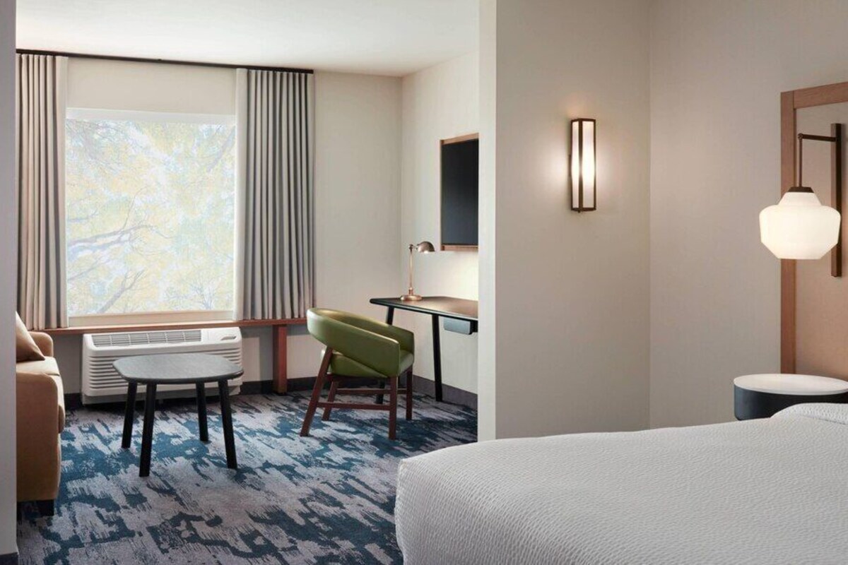 Une chambre d’hôtel avec un lit, un bureau, une chaise, une lampe et une grande fenêtre avec des rideaux.