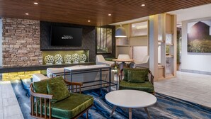 A modern hotel lobby with a stone wall, a flat-screen TV, a seating area with green upholstered chairs, a round table, and a blue patterned carpet.