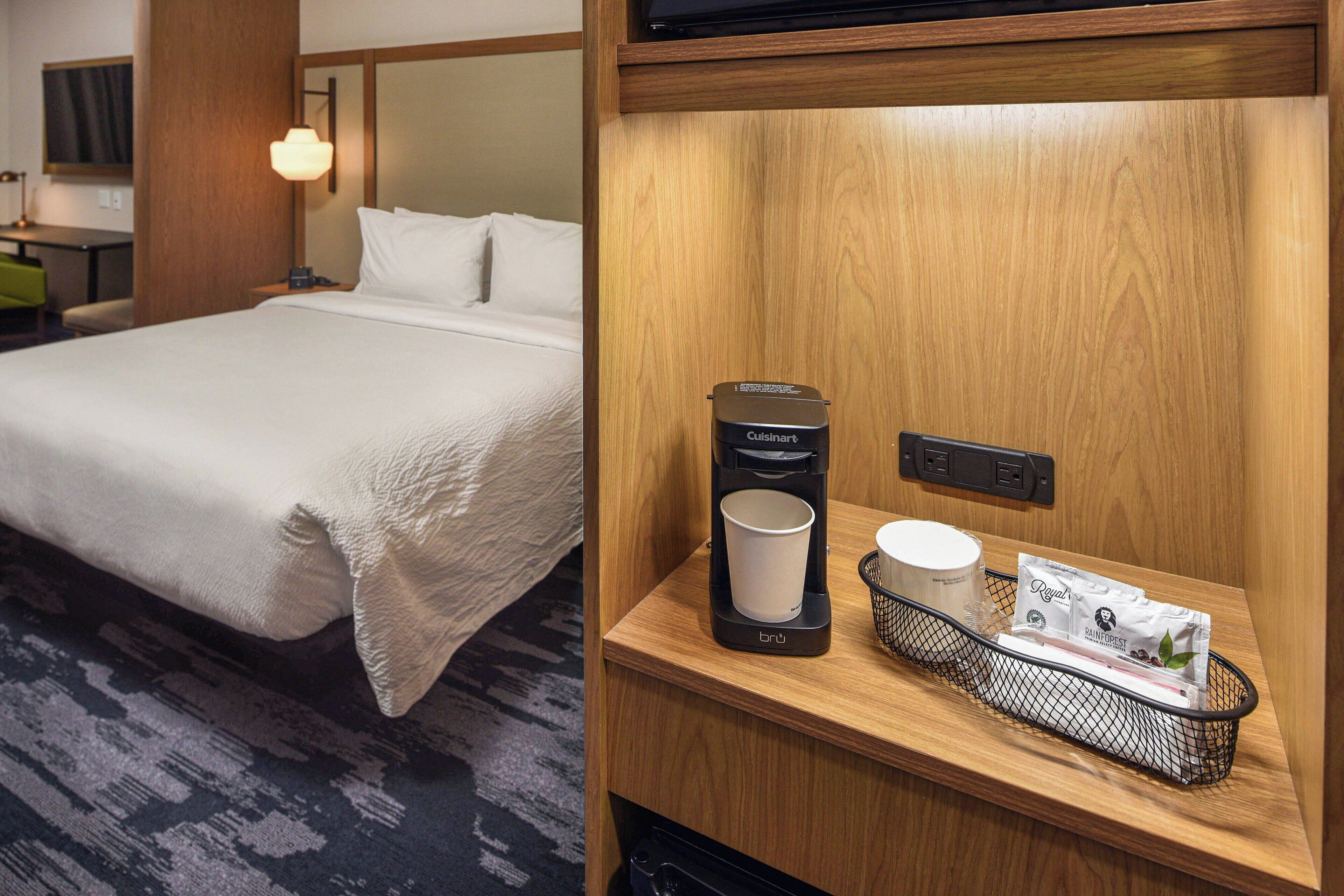 Habitación de hotel con una cama bien hecha, una mesita de noche con una cafetera, una taza y una bandeja con sobres de café.
