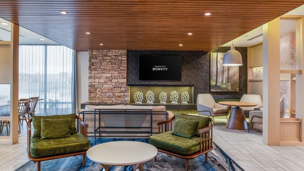 A modern hotel lobby with a stone accent wall, a flat-screen TV displaying 'Marriott Bonvoy', and a seating area with green upholstered chairs and a round table.