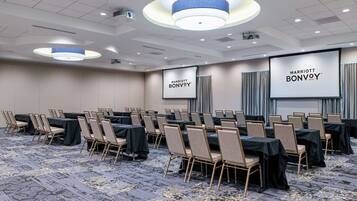 Uma sala de conferências com fileiras de mesas e cadeiras, uma tela grande exibindo 'Marriott Bonvoy' e um design de teto moderno.