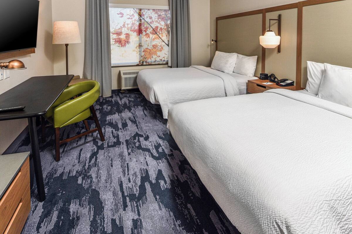 Ein Hotelzimmer mit zwei Betten, einem Schreibtisch mit Stuhl, einem Fernseher und einem Fenster mit Vorhängen.