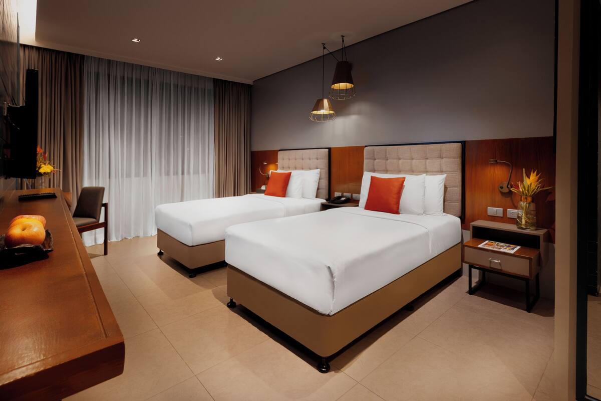 A hotel room with two beds, a desk with a lamp, and a nightstand with a vase.
