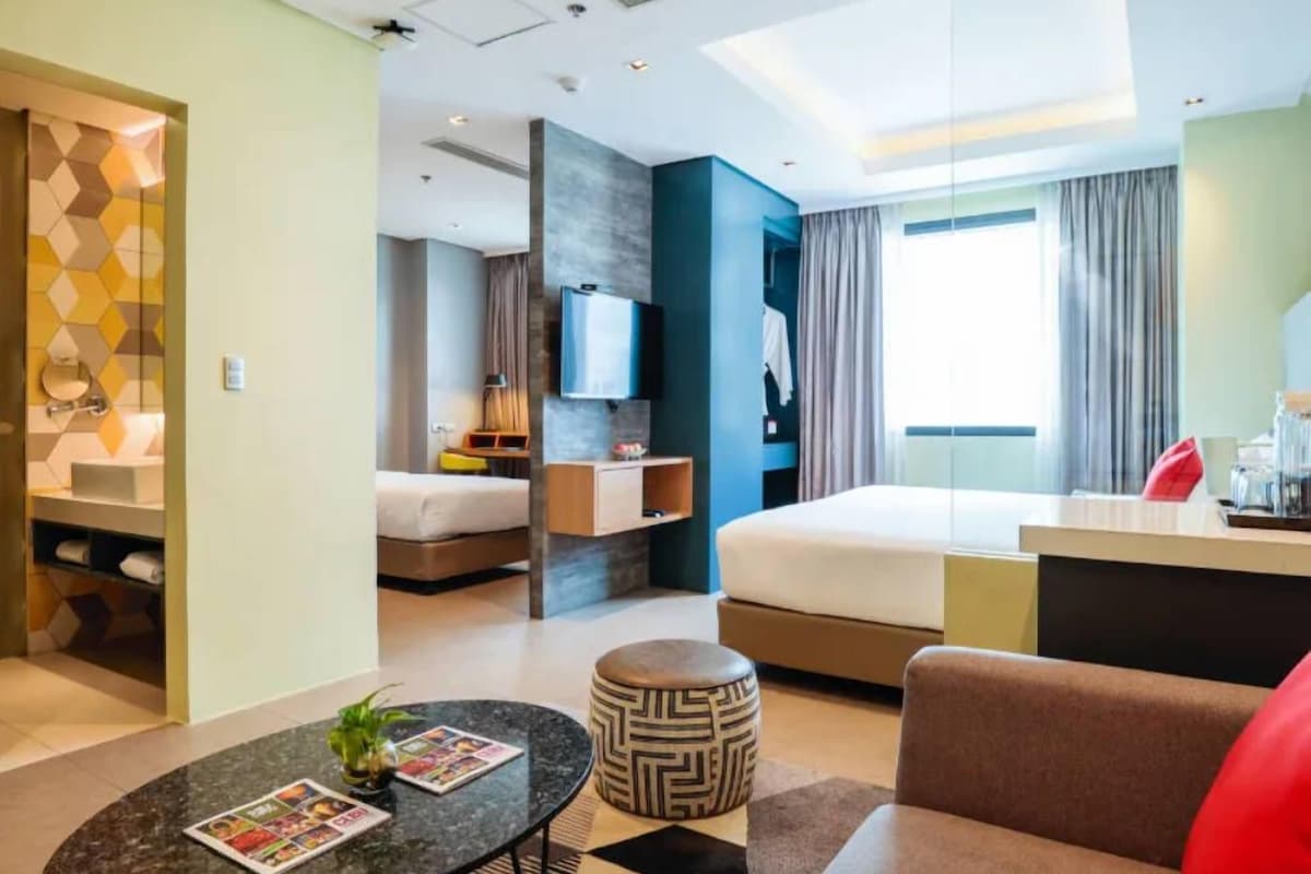 A modern hotel room with a bed, a desk, a TV, and a bathroom visible through an open door.