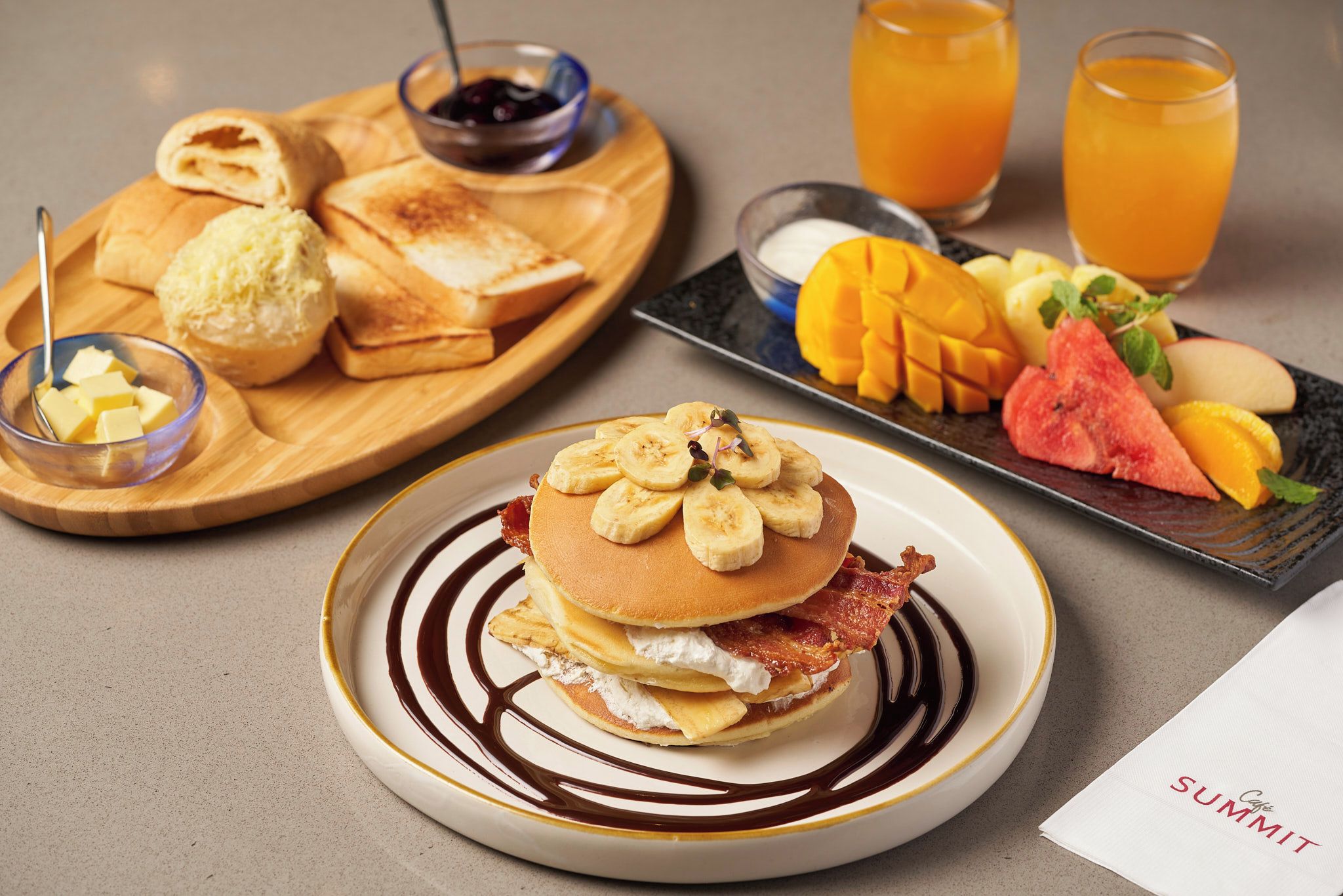 A breakfast plate with pancakes, bacon, and bananas, accompanied by toast, butter, and a fruit plate.