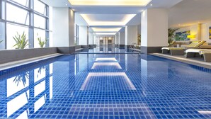 Una gran piscina interior con paredes de vidrio transparente, suelo de baldosas y una zona de estar con cojines y una lámpara.