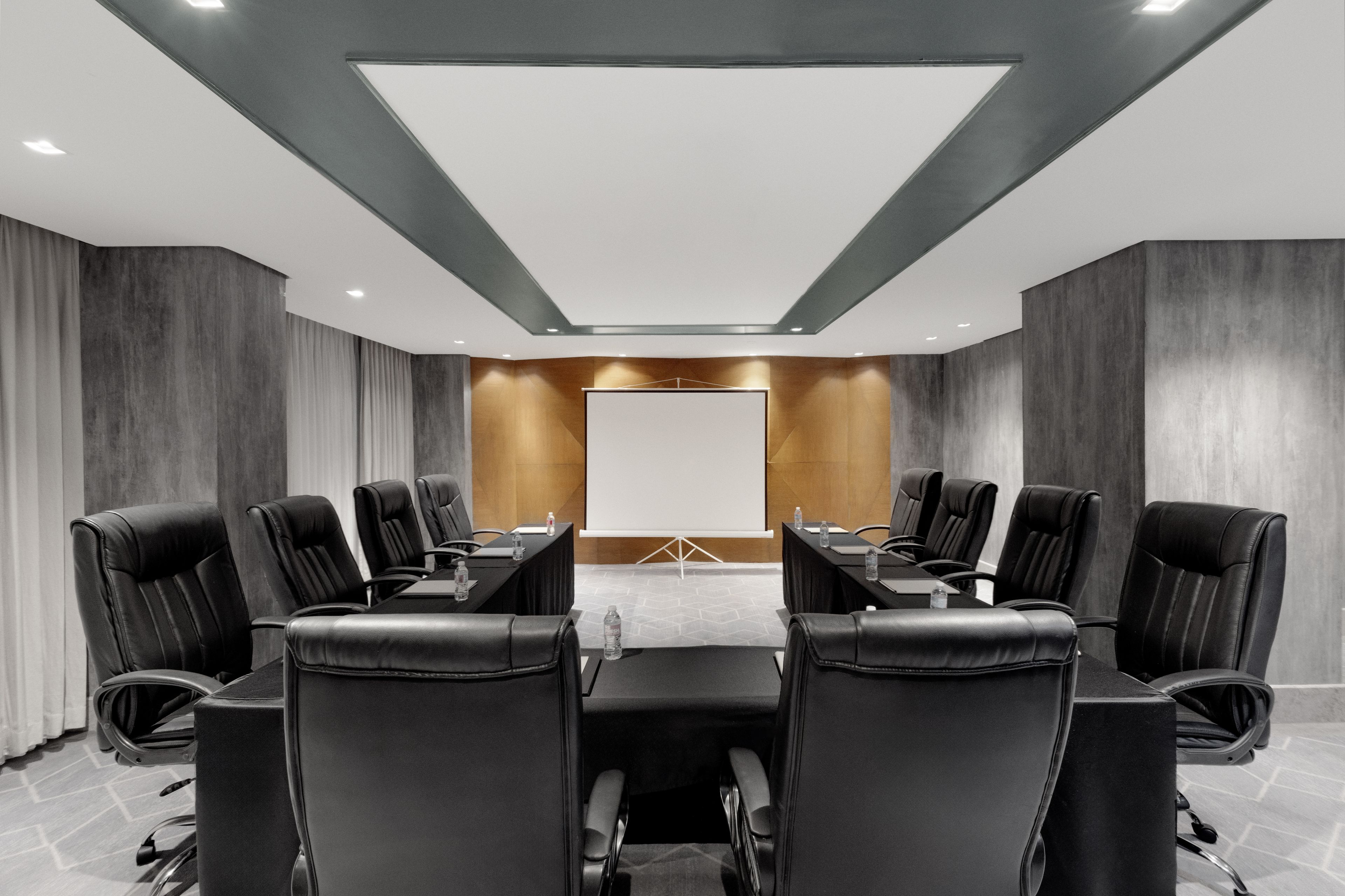 A conference room with a long table, black leather chairs, a projector screen, and a modern ceiling design.