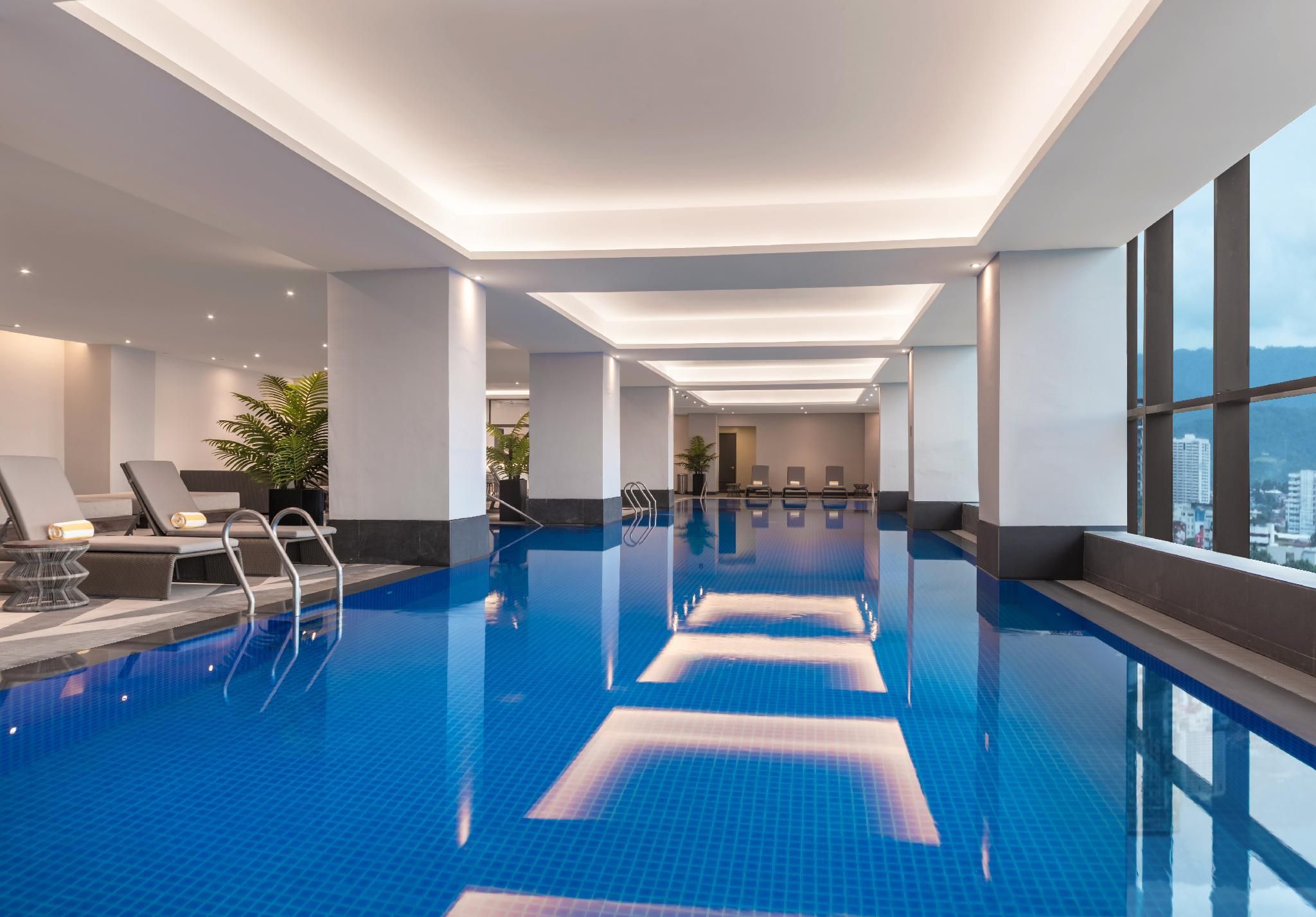 A modern indoor swimming pool with clear blue water, sunlit ceiling, and a view of the city through large windows.