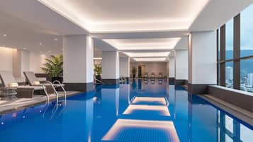 A modern indoor swimming pool with clear blue water, sunlit ceiling, and a view of the city through large windows.
