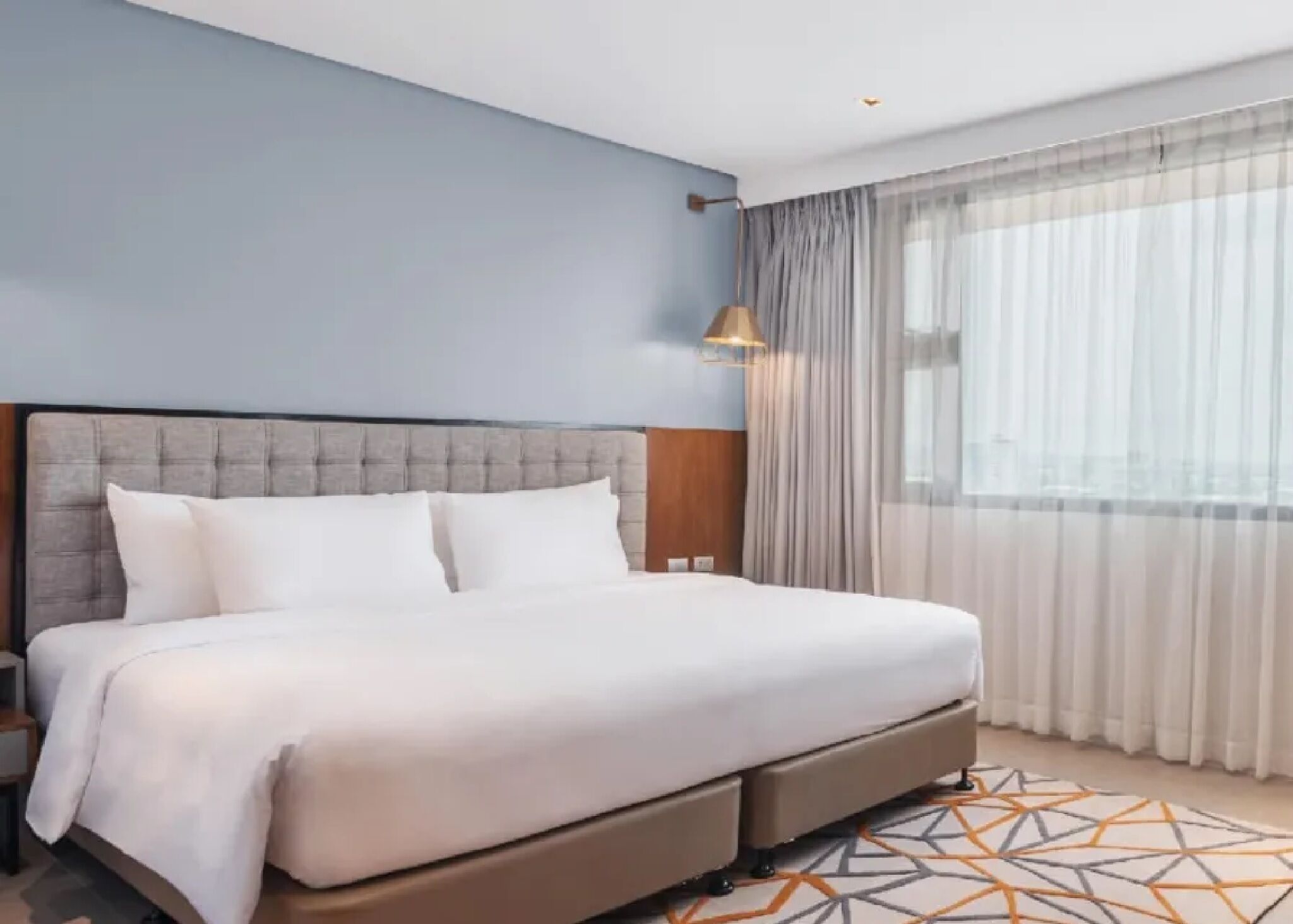 A modern hotel room with a large bed, a nightstand, a lamp, and a view of the city through sheer curtains.