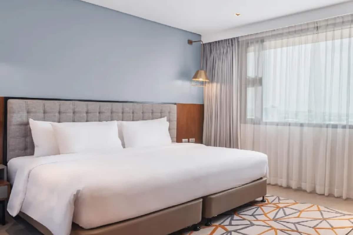 A modern hotel room with a large bed, a nightstand, a lamp, and a window with sheer curtains.